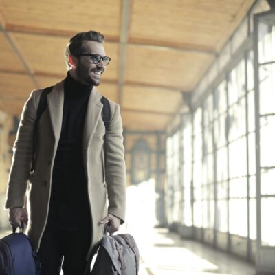 Smiling man in a winter coat with bags walking through a bright train station corridor.