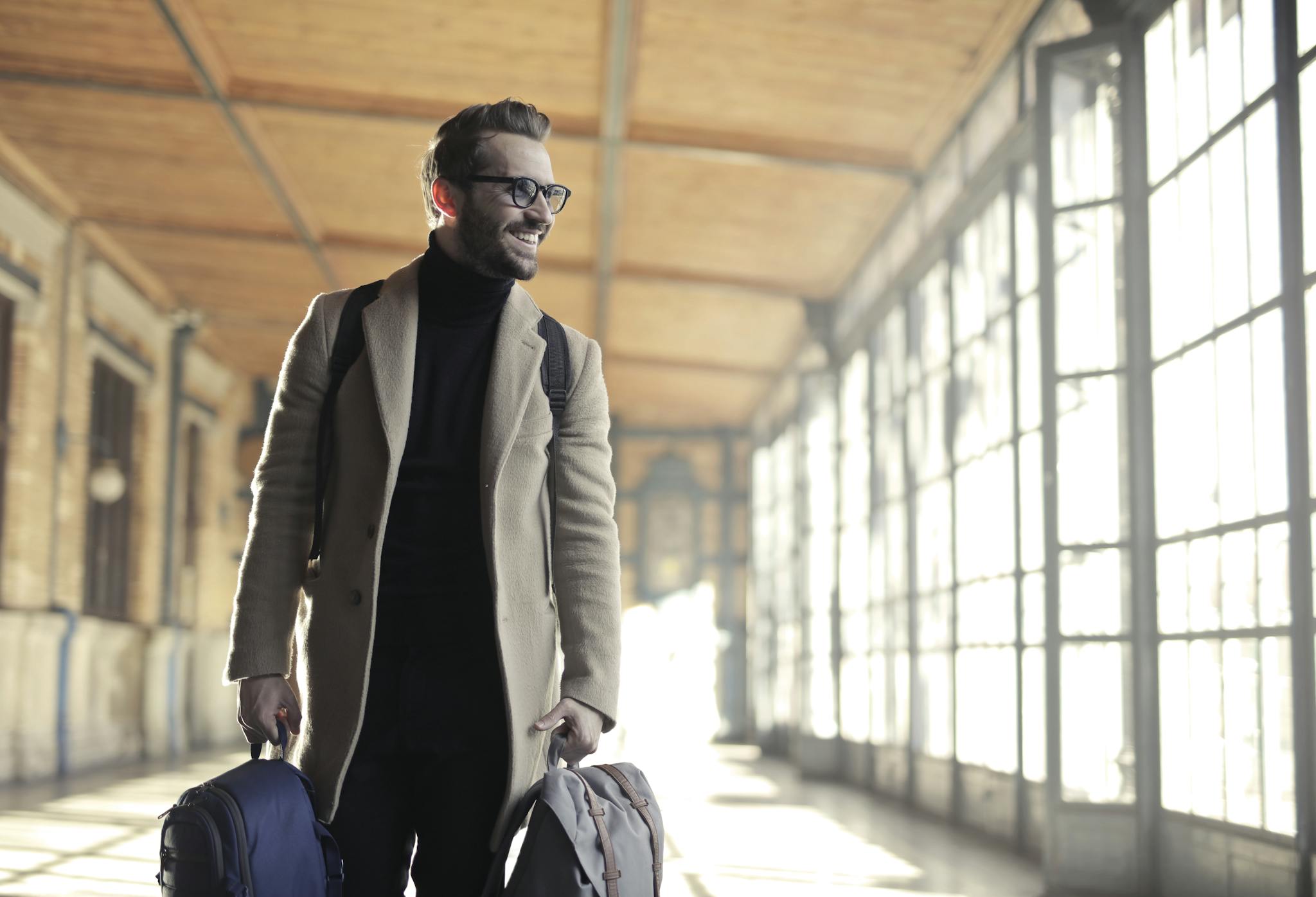 Smiling man in a winter coat with bags walking through a bright train station corridor.